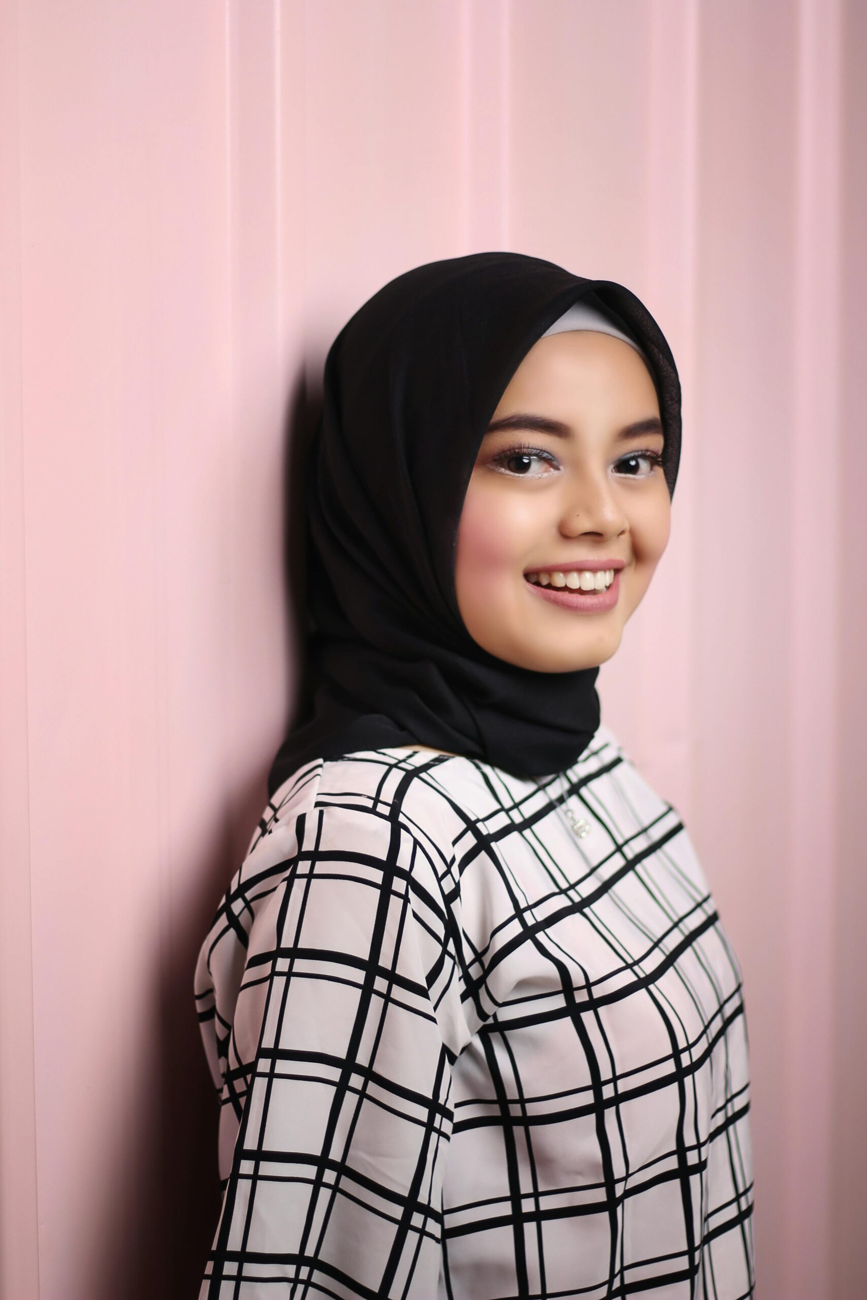 A young woman wearing a hijab smiles warmly against a pastel pink background.
